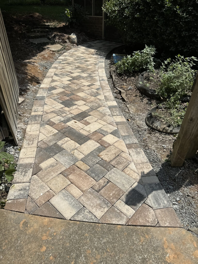 A newly installed stone paver walkway with a herringbone pattern curves through a garden area bordered by plants and gravel.