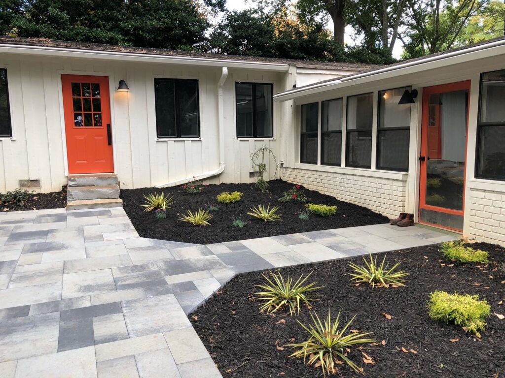 Single-story white house with orange doors, a paved walkway, and mulched garden beds featuring small green plants in the front yard.