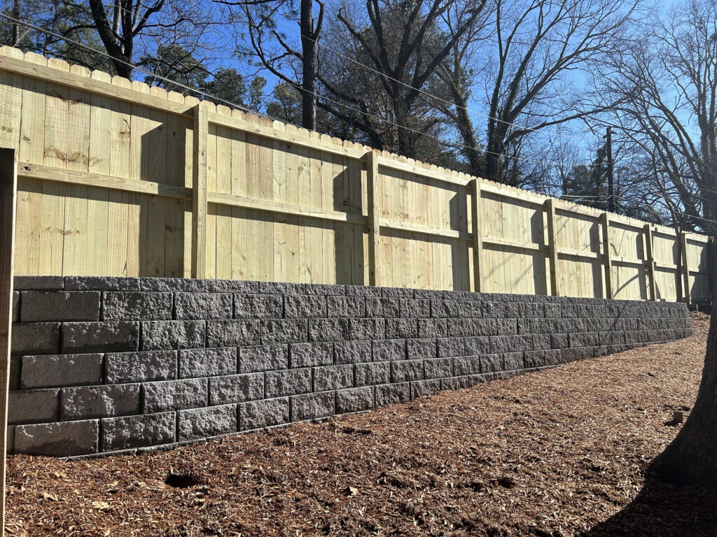 A wooden privacy fence stands atop a gray concrete retaining wall, with mulch covering the ground and bare trees visible in the background.