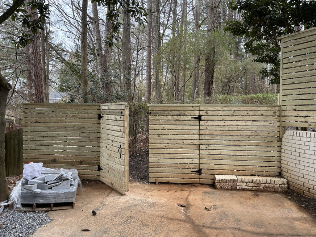 A wooden gate is open in a horizontal slat fence, with a stack of construction materials and bricks on the concrete ground. Trees and woods are visible in the background.