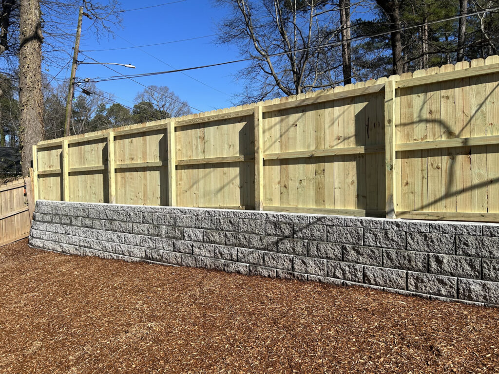 A wooden privacy fence sits atop a gray stone retaining wall, with wood chips covering the ground and leafless trees in the background.
