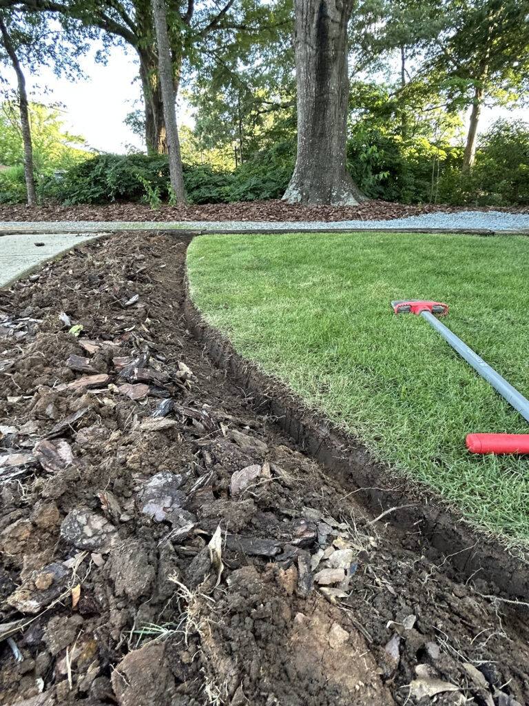 A freshly dug trench separates a patch of grass from a mulched area, with a red garden tool lying on the lawn beside the trench.