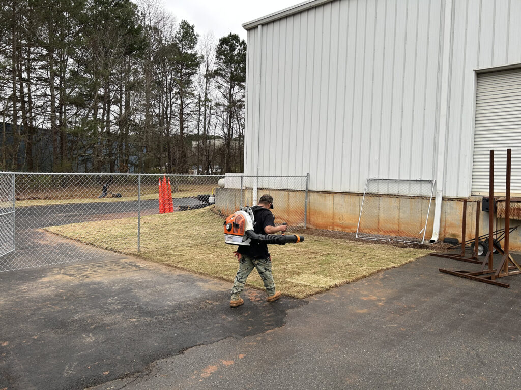 A person wearing a leaf blower walks on a paved area next to a fenced, grassy patch beside a large industrial building.