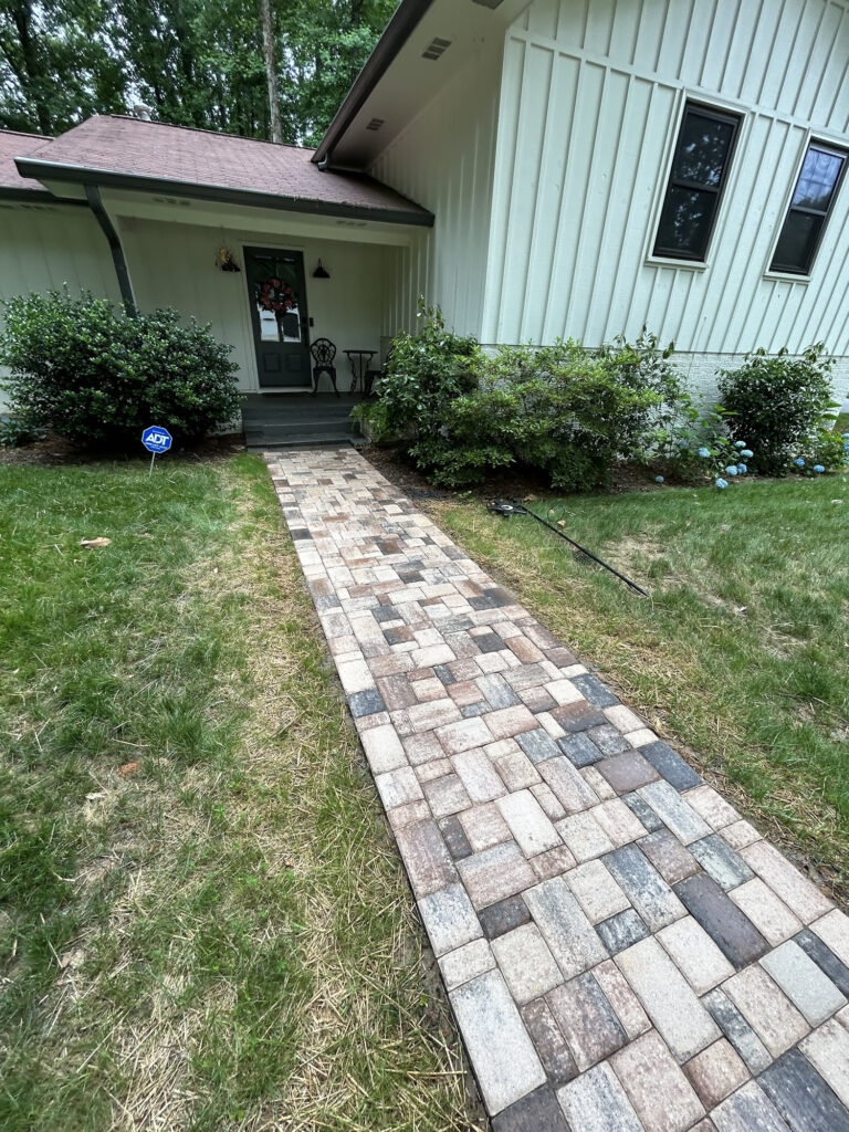 A paved stone walkway leads to the front door of a house with white siding, flanked by bushes and a patch of grass on each side.