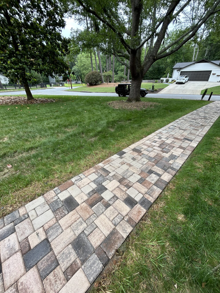 A paved walkway made of rectangular bricks runs through a grassy yard toward a street with houses, trees, and a parked black truck.