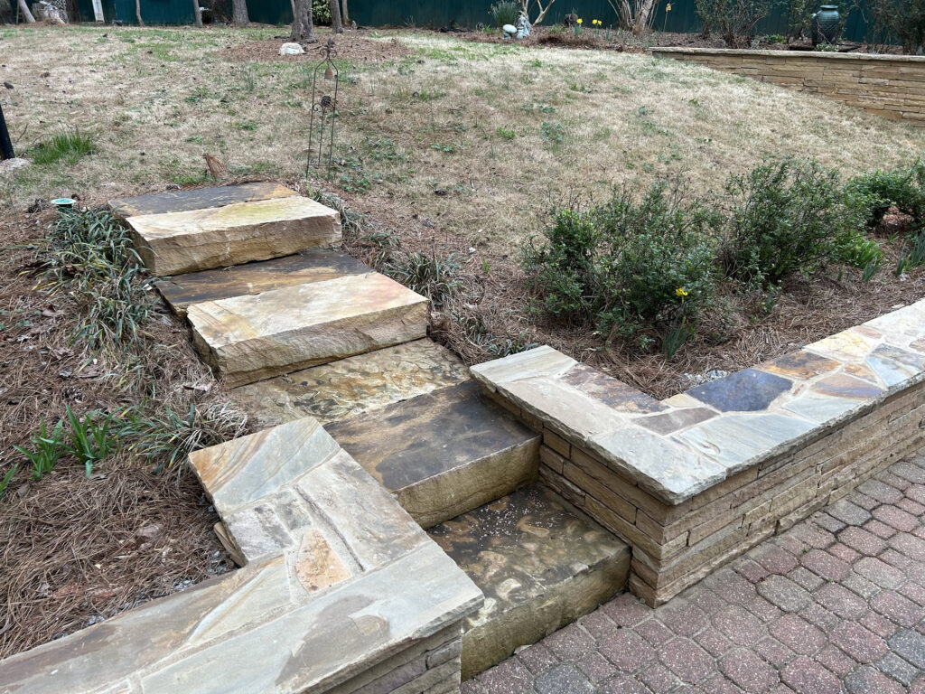 Stone steps lead from a paved area up to a grassy yard, bordered by low stone walls and some small shrubs.