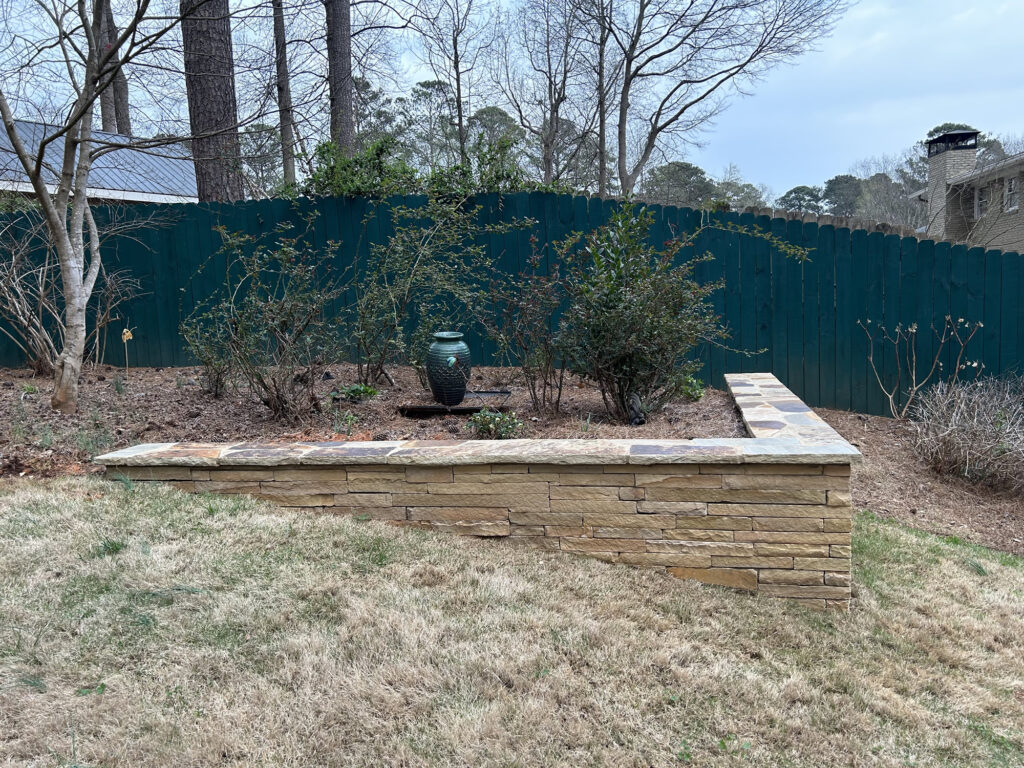 A stone retaining wall encloses a small garden bed with bushes and a green ceramic vase, set in a grassy yard with a teal wooden fence and trees in the background.