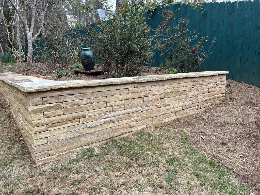 A stacked stone retaining wall borders a garden bed with shrubs and a large green pottery urn, beside a green wooden fence.