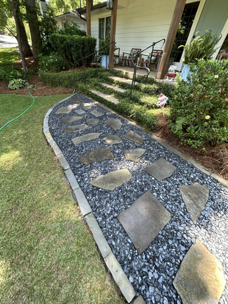 Stone pavers set in black gravel form a walkway leading to a porch with steps, surrounded by green shrubs and a manicured lawn.