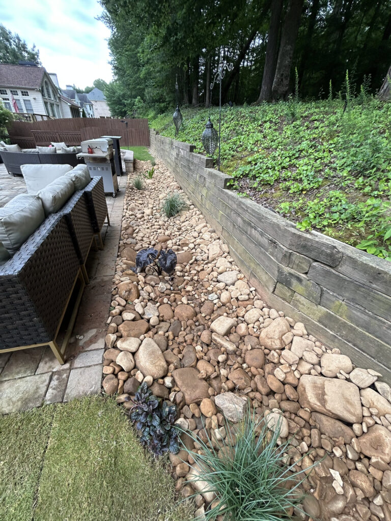 A dry creek bed made of various sized rocks runs alongside a patio with outdoor furniture, bordered by a wooden retaining wall and a sloped, vegetated area.