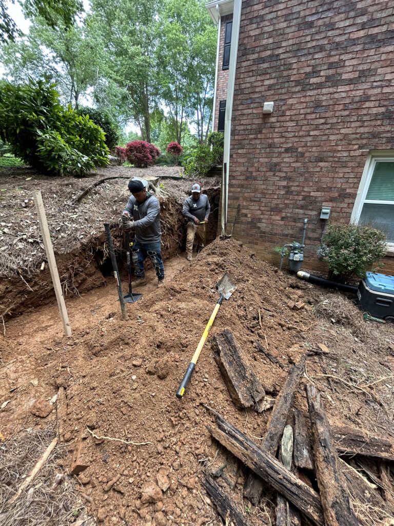Two workers dig a trench next to a brick house, surrounded by dirt, tools, and exposed tree roots. Shrubs and trees are visible in the background.