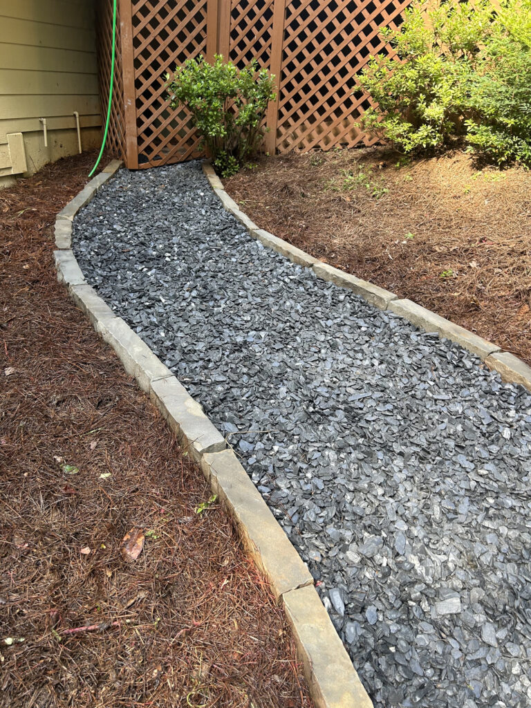 A gravel path bordered by stone pavers leads to a wooden lattice gate, with a green garden hose visible along the house wall and shrubs on both sides.