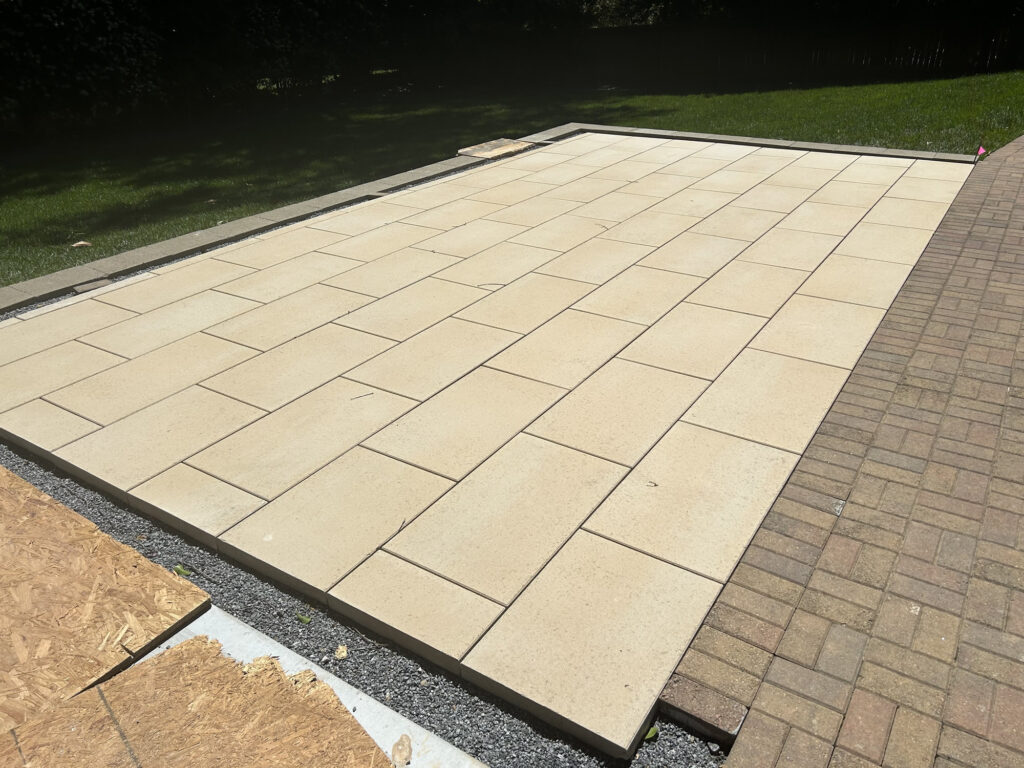 A newly installed rectangular patio with light-colored rectangular tiles next to an existing brick patio, bordered by grass and construction materials.