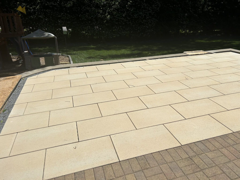 A patio area with large rectangular beige pavers arranged in a grid, next to a lawn and bordered by smaller brown bricks.