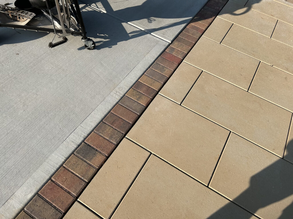 Concrete sidewalk with a red brick border next to tan paving stones; partial shadow and construction equipment visible.