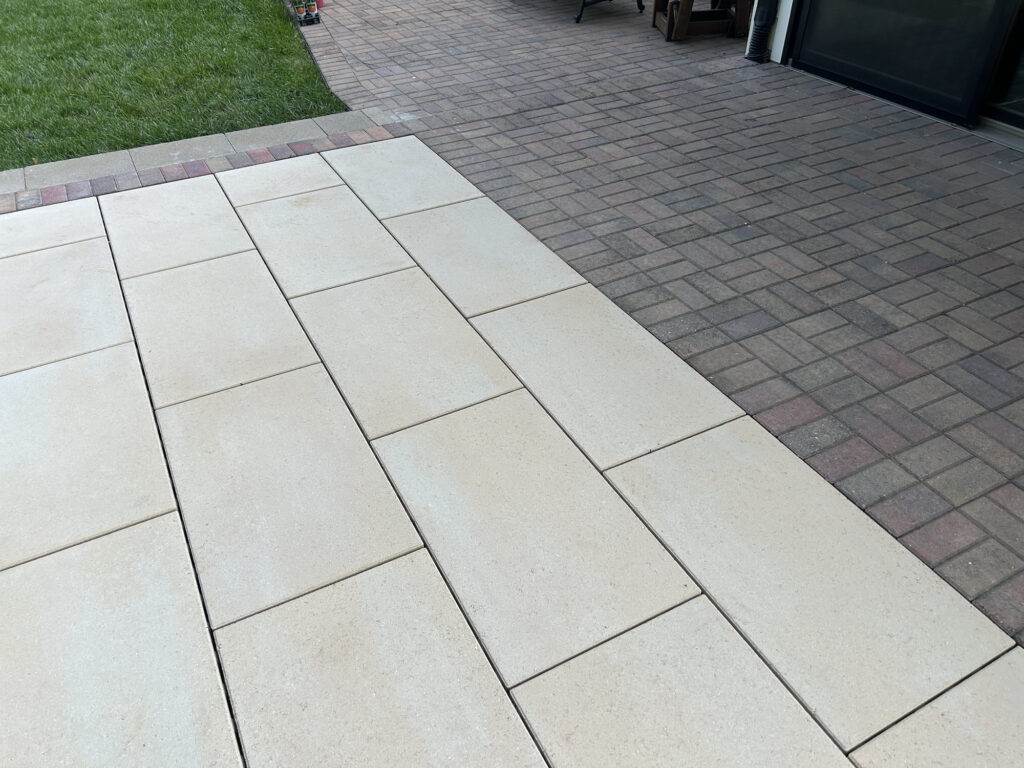 A patio with large rectangular light-colored pavers adjacent to an area with smaller, darker square pavers, bordered by green grass.