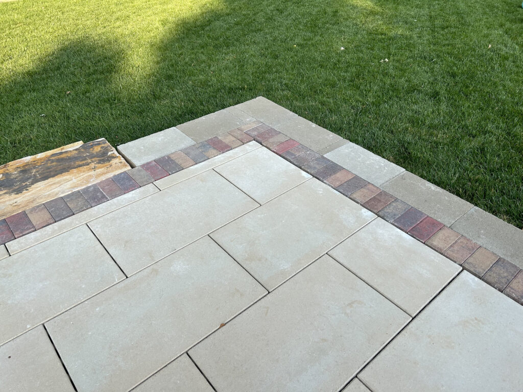 A patio made of large rectangular pavers bordered by red and brown bricks meets a green lawn.