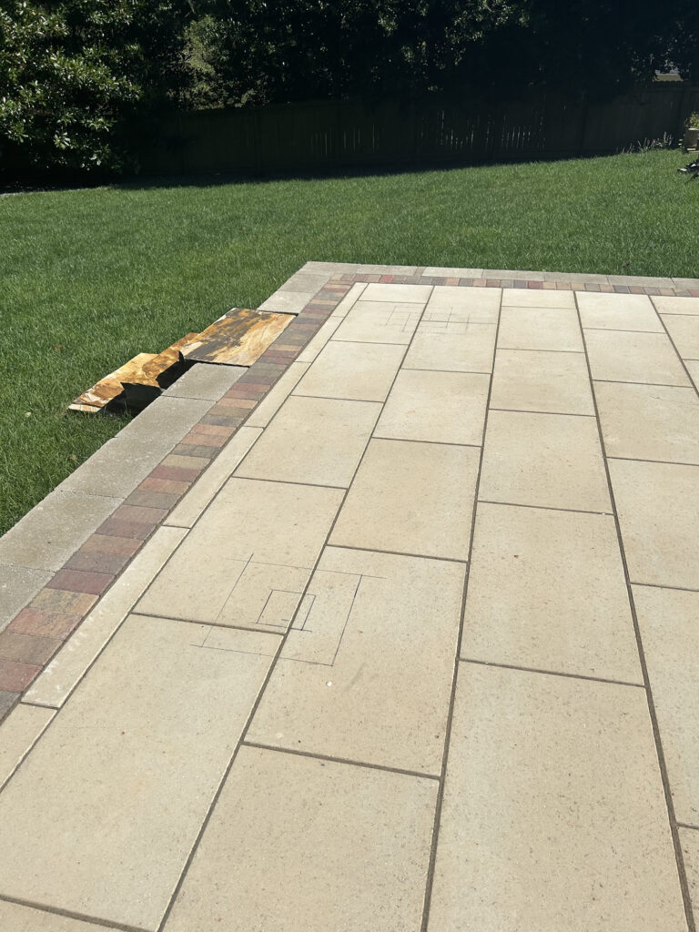 A paved patio with rectangular tiles, some with sketched outlines. Brown tarps or boards lie on the grass beside the patio, with a lawn and trees in the background.