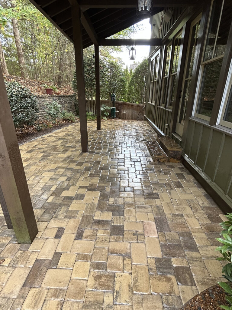 A covered patio with wet tan and brown stone pavers, next to a green house with large windows, surrounded by trees and shrubs.