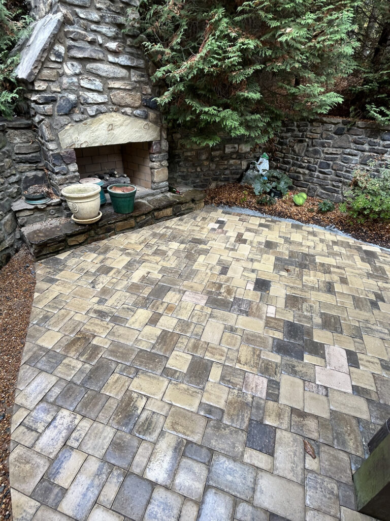 Stone patio with a variety of beige and gray pavers, an outdoor stone fireplace, potted plants, and a stone wall surrounded by greenery.