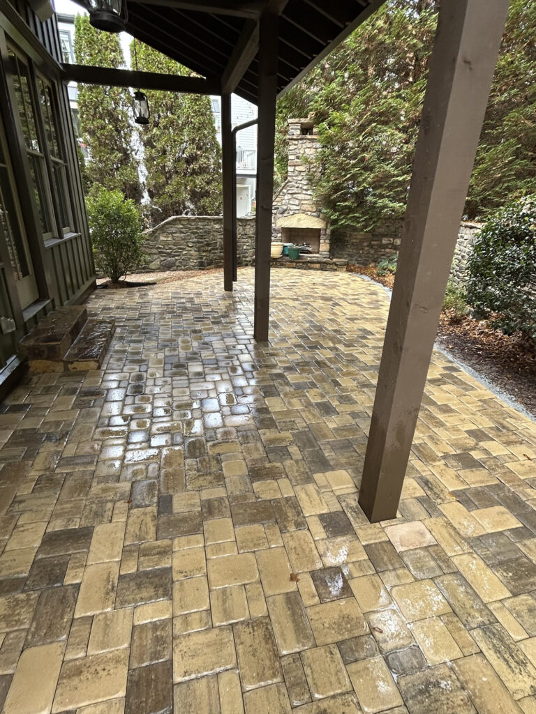 A stone patio with a wooden pergola, bordered by shrubs and trees, featuring a built-in outdoor stone fireplace at the far end.