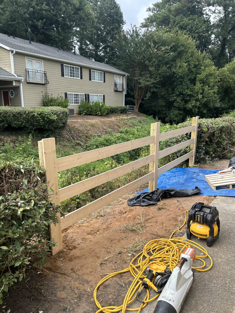 Partially constructed wooden fence with power tools and extension cords on the ground in front of a house with green bushes and trees in the background.