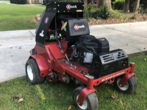 A red Exmark lawn aerator machine is positioned on grass near a concrete driveway, with trees and a lawn in the background.