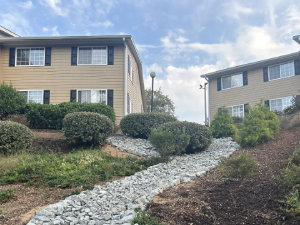 Two beige apartment buildings with black shutters and white trim stand on either side of a rocky pathway, surrounded by bushes and greenery under a partly cloudy sky.
