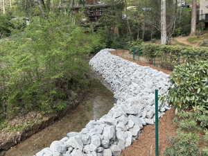 A small creek bordered by a bank of stacked white rocks, with greenery and trees on both sides and a building visible in the background.