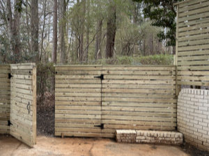 Wooden fence with horizontal slats and a gate partially open, set on a concrete surface with trees and shrubs in the background.