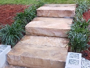 Three large rectangular stone steps bordered by green plants and mulch in a landscaped outdoor area.