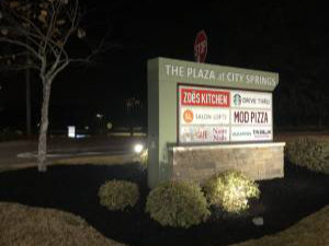 Illuminated sign for The Plaza at City Springs at night, displaying logos for various restaurants including Zoës Kitchen, MOD Pizza, and others.