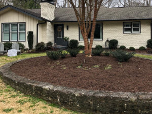 A mulched circular flower bed with small shrubs in front of a single-story house with cream-colored brick siding.