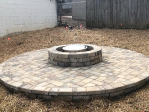 Circular stone patio with a raised stone fire pit in the center, surrounded by dry grass and enclosed by wooden and concrete fences.