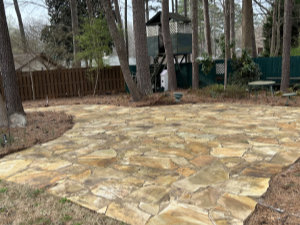 A stone patio is shown in a backyard with tall trees, a wooden fence, a play structure, and a bench in the background.