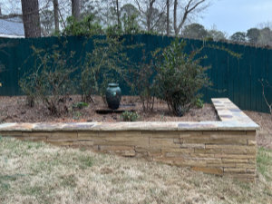 Raised stone garden bed with sparse shrubs and a green urn in the center, set against a green wooden fence and background trees.