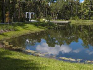 A small pond with grassy banks reflects trees and clouds, with a paved path and a white, rectangular structure visible in the background.