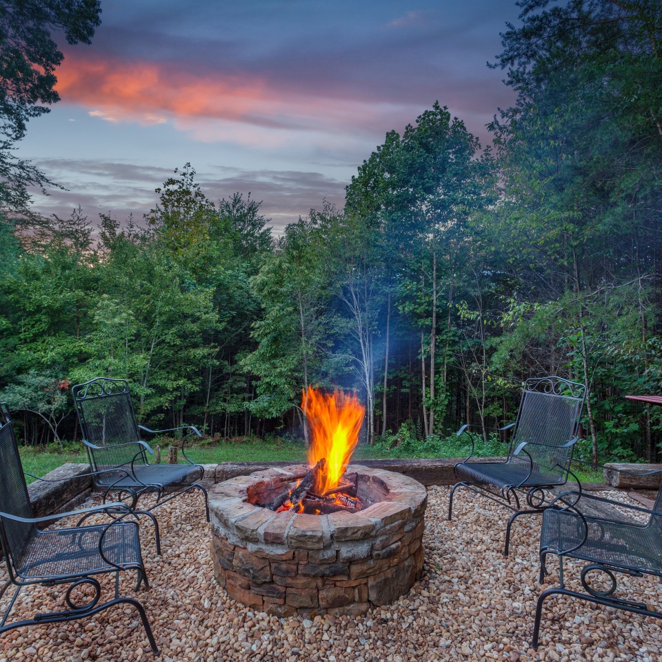 Roaring fire in a stone firepit with wood logs and surrounded by trees at dusk in a backyard