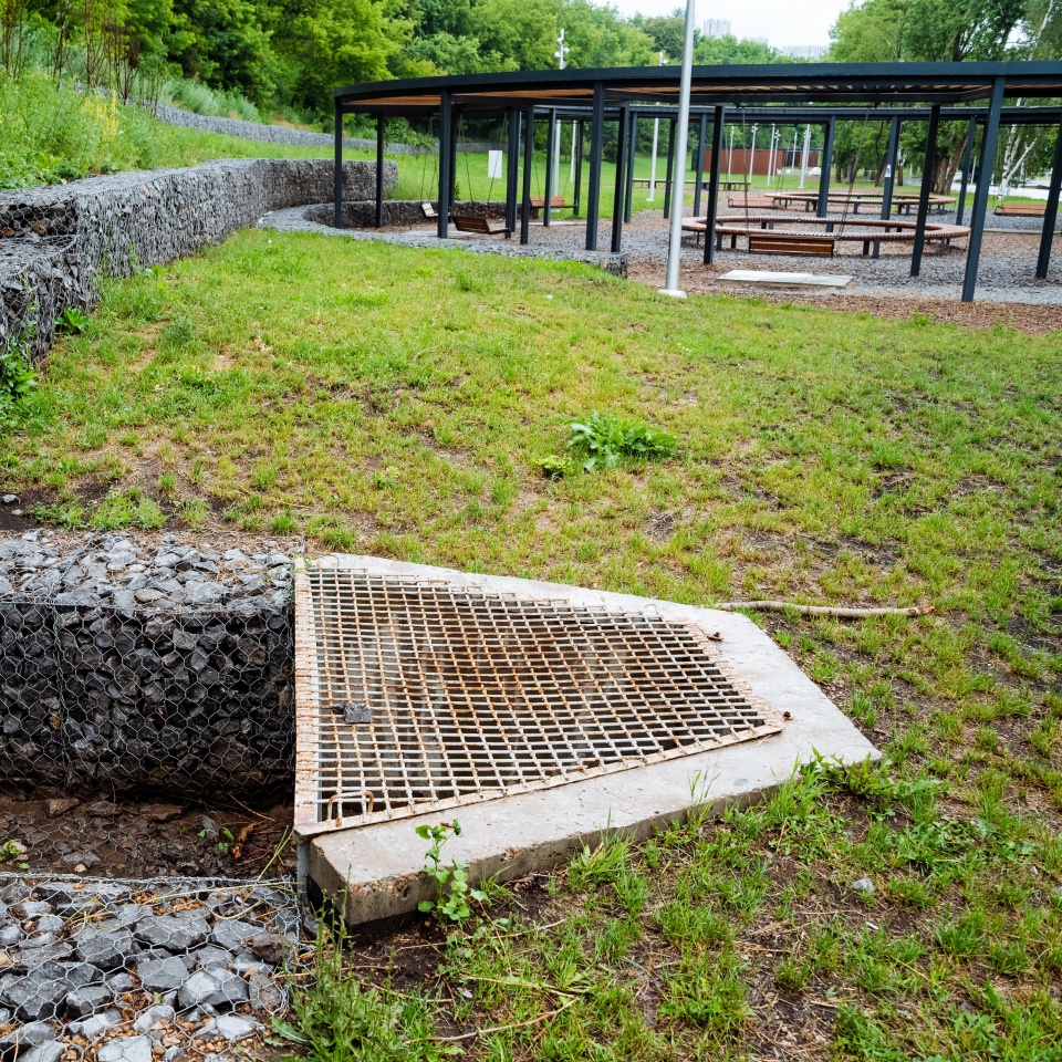 Outdoor drainage system featuring gravel, stone steps, and a metal grate. The system is part of a modern urban park with walking paths and green spaces, showcasing effective water management.