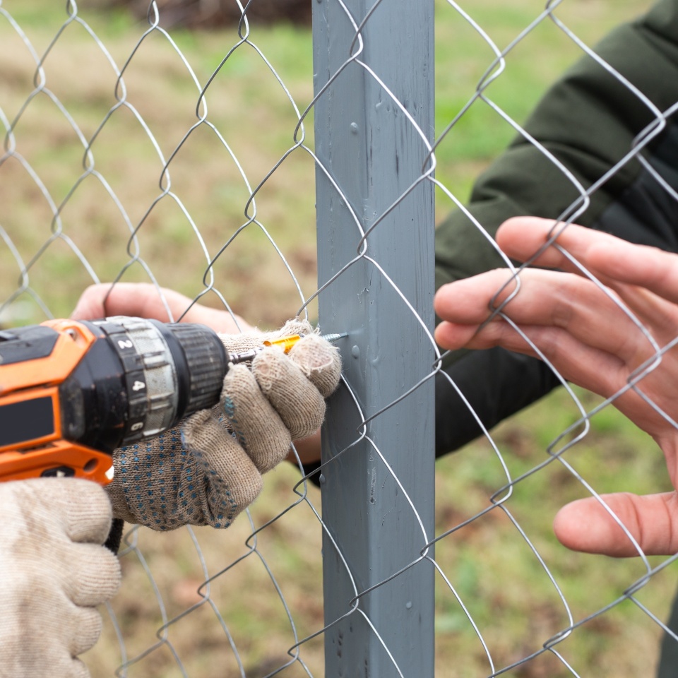 A man is building a fence at his dacha. Using an electric drill, he screws a metal mesh chain-link to the metal profile.