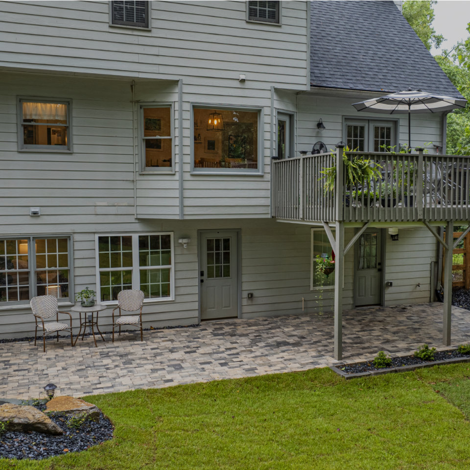 Back view of a light gray, two-story house with a raised deck, patio seating area, paved ground, and green lawn.