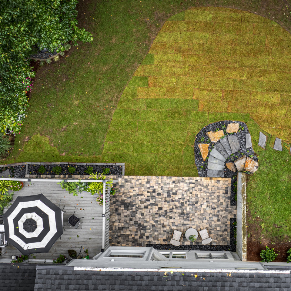 Aerial view of a backyard with a stone patio, a black-and-white umbrella, and newly laid sod next to a curved stone walkway and steps.